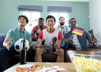 88,2% dos brasileiros avaliam positivamente empresas que flexibilizam a rotina durante jogos da Copa do Mundo 2 multiracial curious male friends with beer bottles flags ball watching soccer match home scaled