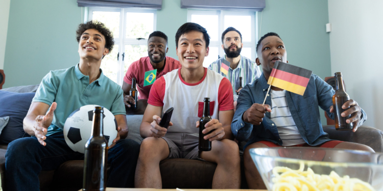 88,2% dos brasileiros avaliam positivamente empresas que flexibilizam a rotina durante jogos da Copa do Mundo 1 multiracial curious male friends with beer bottles flags ball watching soccer match home scaled
